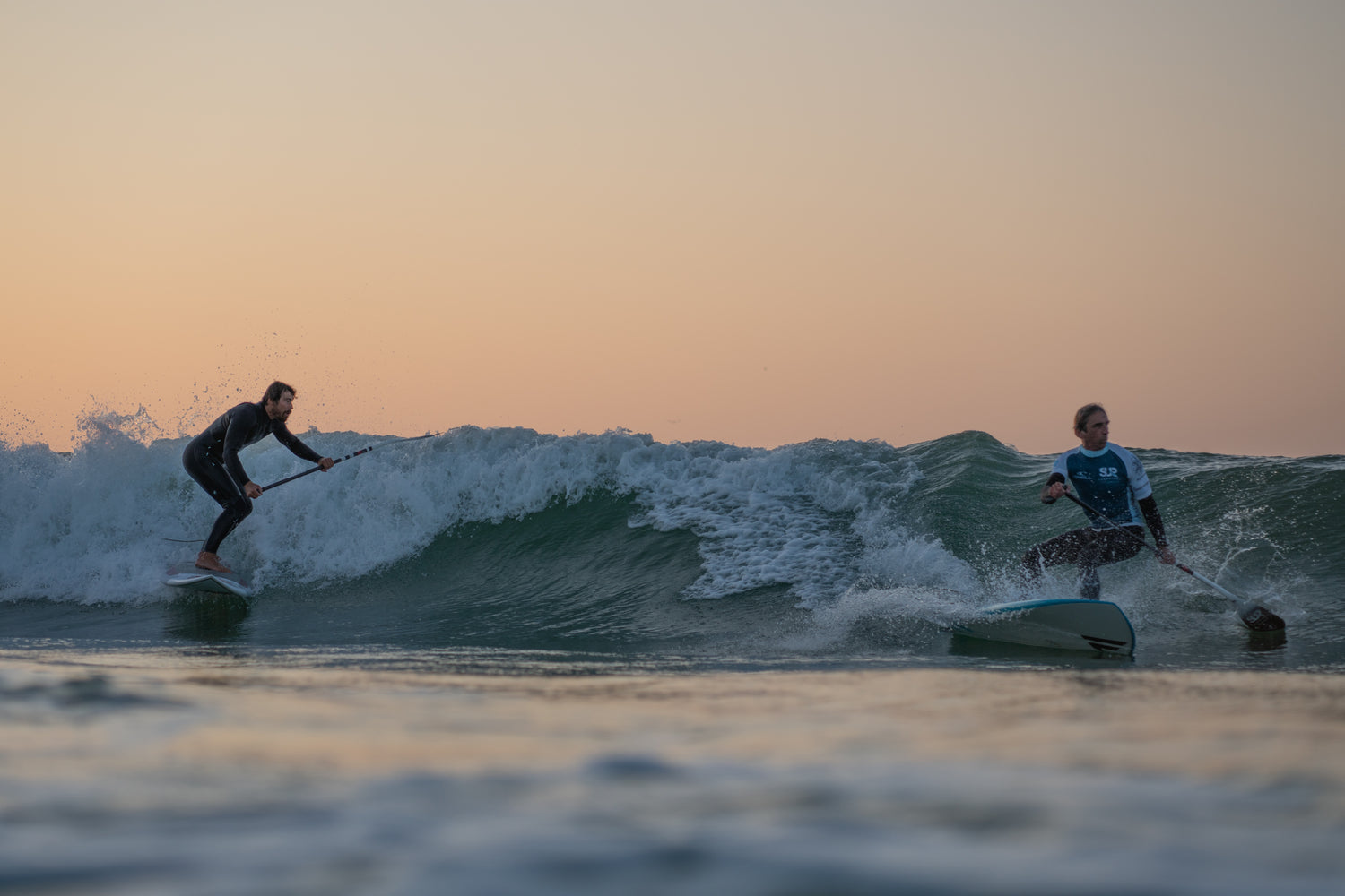 Kai Steimer und Thomas Wendt teilen sich im Sonnenuntergang eine Welle beim Wave SUP Surfen.