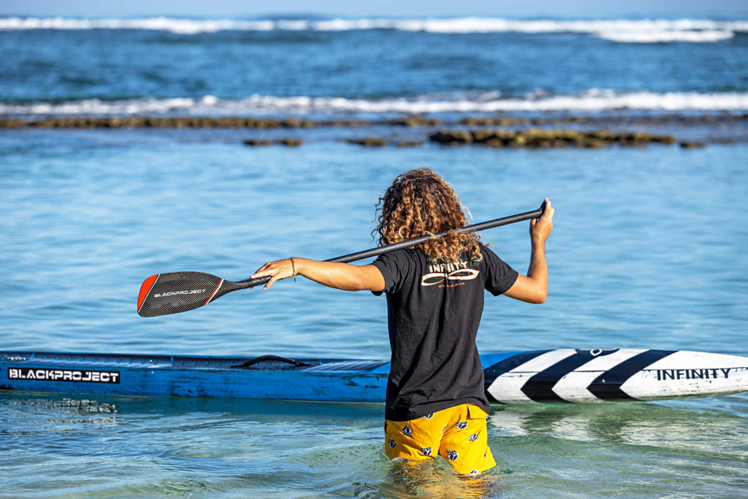 Stand up Paddler mit einem Black Project TempoX SUP Race Touring Paddel quer über den Schultern im hüfttiefen Wasser.