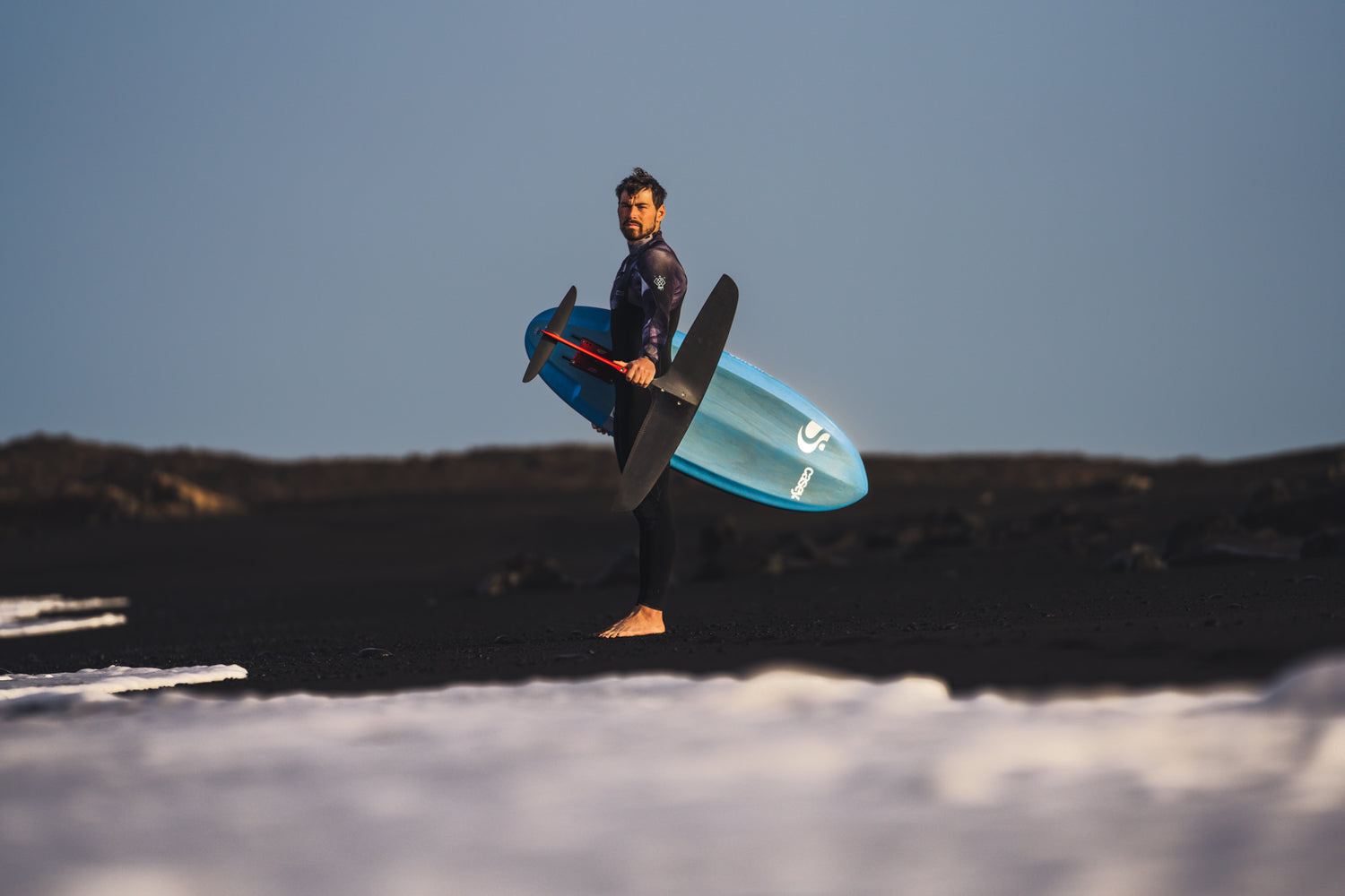 Prone Foiler Kai-Nicolas Steimer posiert am Strand mit dem Sunova Casey Pilot Surf Foil Board und einem Axis PNG Foil Set up.